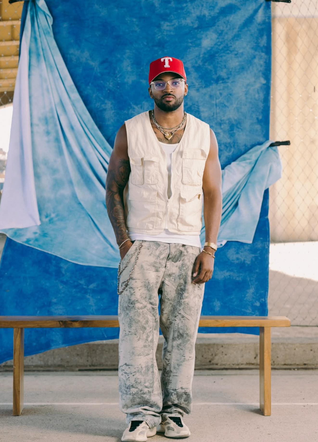 Kingdom Jones standing full-body in front of a blue backdrop wearing a red cap and cream vest